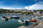 Harbor scene with boats docked along a rocky shore, houses in the background under a blue sky.